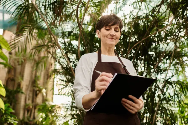 Mature woman joyfully manages her tasks in a lively greenhouse surrounded by plants. — Stock Photo
