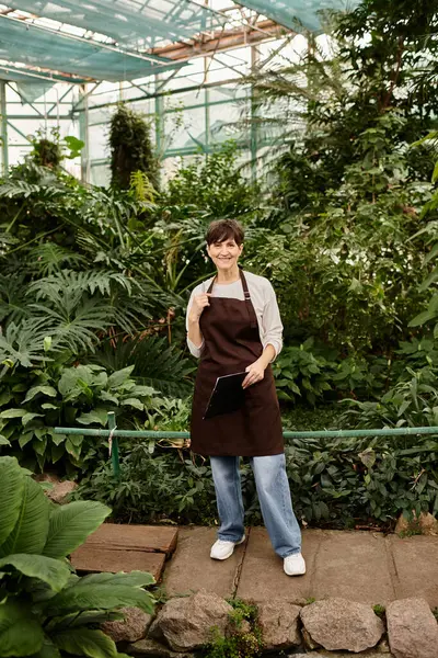 Confident mature woman joyfully works among lush greenery in a well maintained greenhouse. — Stock Photo
