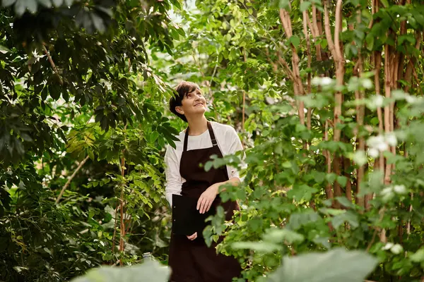 Mujer madura con sonrisas de pelo corto mientras trabaja entre plantas prósperas en un invernadero. - foto de stock