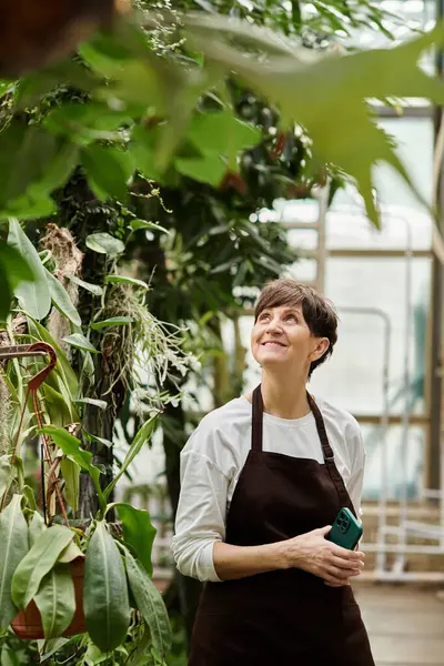 Confident woman in a greenhouse admires the thriving plants while holding her phone in hand. — Stock Photo