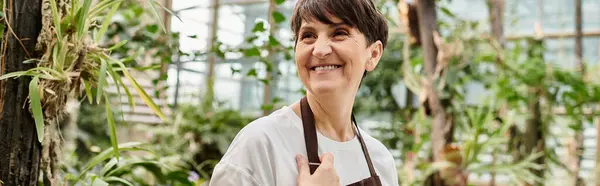 Mujer madura con el pelo corto sonríe con confianza mientras que tiende a las plantas en un animado invernadero. - foto de stock