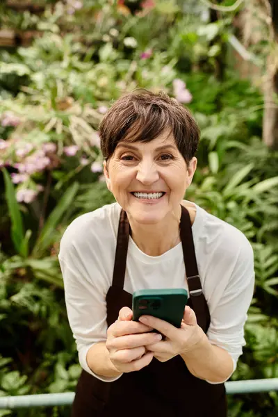 A confident woman smiles while using her phone among flourishing plants in a beautiful greenhouse. — Stock Photo