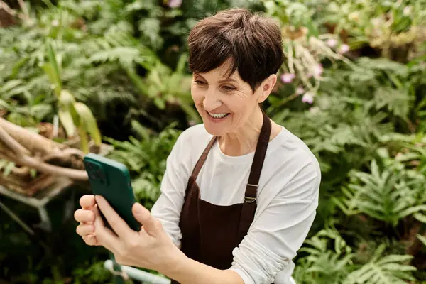 Mature woman smiles while using her smartphone, surrounded by vibrant greenery in a greenhouse. — Stock Photo