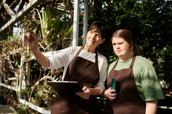 A mature woman guides her young colleague with Down syndrome in a lush greenhouse setting. — Stock Photo