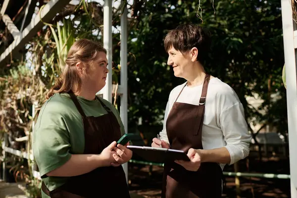 Mature woman supports her young colleague with Down syndrome while working in a vibrant greenhouse. — Stock Photo