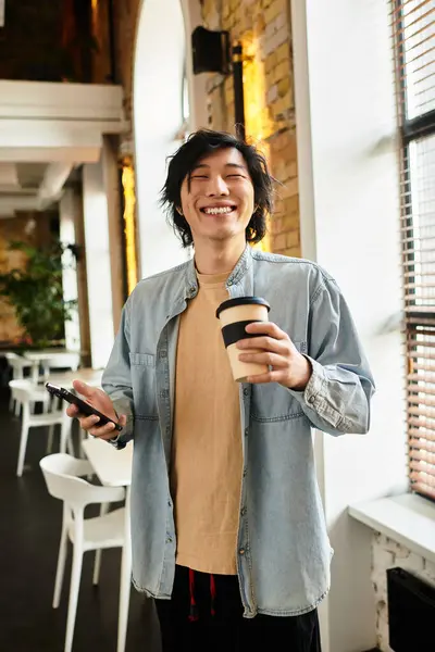 Young man smiles broadly while holding a coffee cup and phone in a lively office environment. — Stock Photo