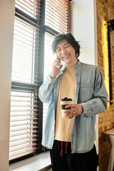 A young individual smiles while talking on the phone, holding a coffee cup in a bright office. — Stock Photo