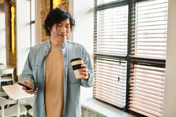 A young man is savoring his coffee, absorbed in his phone, in a stylish office with large windows. — Stock Photo