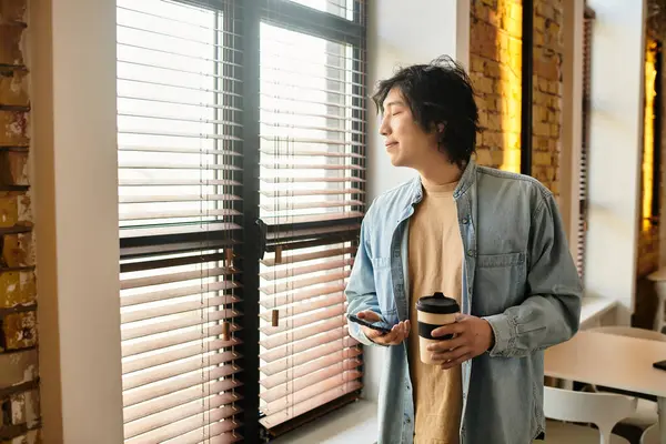 A young person by the window sips coffee and checks their phone, savoring the view. — Stock Photo