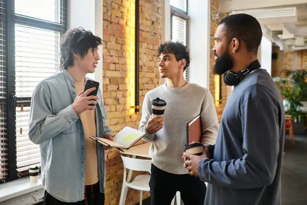 Tre colleghi si impegnano in una discussione animata mentre tengono tazze di caffè in uno spazio di lavoro alla moda. — Foto stock