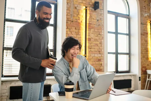 Two coworkers share a light moment over coffee while working on a laptop in a stylish office. — Stock Photo