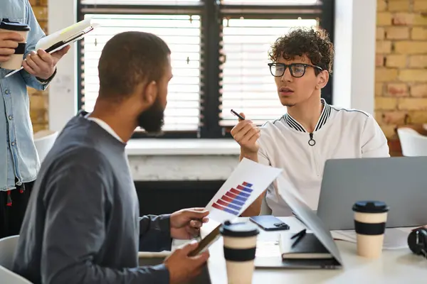 Dois colegas discutem gráficos de projetos enquanto outros assistem e tomam notas. — Fotografia de Stock