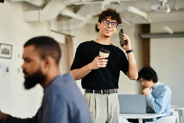 Un hombre disfruta de su café mientras habla por teléfono, rodeado de colegas en un espacio de trabajo luminoso. - foto de stock