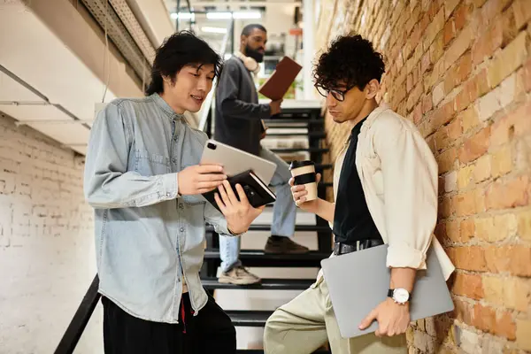 Dos compañeros de trabajo discuten ideas con dispositivos digitales, mientras que otra persona observa desde las escaleras. — Stock Photo