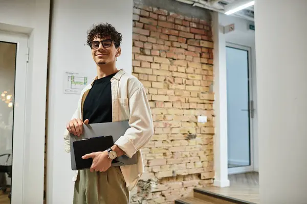 A man with curly hair holds a laptop and notebook while smiling in a stylish workspace. — Stock Photo