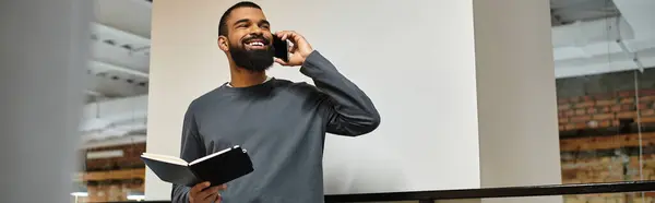 Un uomo sorridente in un ufficio moderno parla al telefono mentre tiene un quaderno, essendo produttivo. — Foto stock