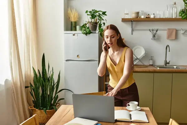 A young woman talks on the phone while getting ready to work at her kitchen table. — Stock Photo