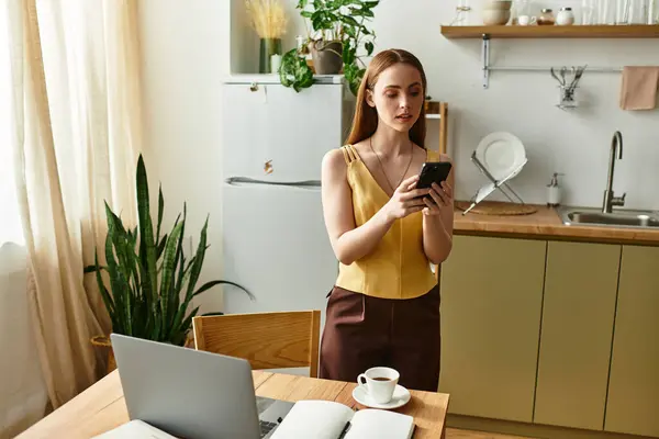 Una bella giovane donna sta accanto a un tavolo di legno, sorseggiando caffè e usando il suo smartphone. — Stock Photo