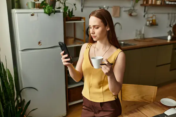 Sunlight streams into the kitchen as a young woman enjoys her coffee and checks her phone. — Stock Photo