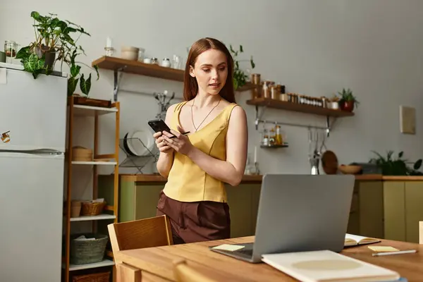 In a bright kitchen, a young woman studies her phone, focused by her laptop. — Stock Photo