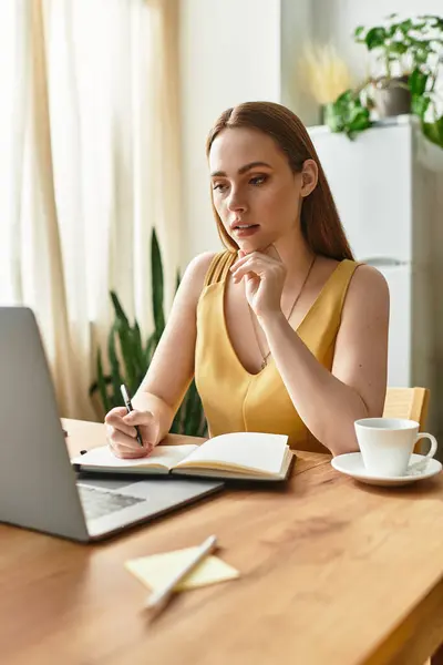 Young woman in a yellow dress thoughtfully jots notes while working on her laptop at home. — Stock Photo
