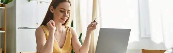 Una joven hermosa mujer sonríe alegremente mientras trabaja en su computadora portátil en una habitación soleada en casa. — Stock Photo