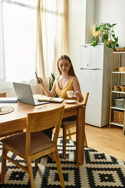 A young woman sits at a table, holding coffee and working on her laptop in a bright room. — Stock Photo