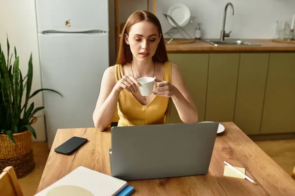 Beautiful young woman sits at a wooden table with her laptop, sipping coffee in a bright kitchen. — Stock Photo