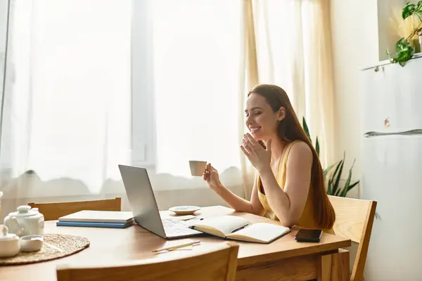 A young woman smiles while sipping tea and working on her laptop in a bright, cozy kitchen. — Stock Photo