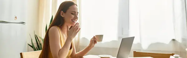 Una giovane donna sorride sorseggiando un caffè e lavorando sul suo portatile in una cucina soleggiata. — Stock Photo