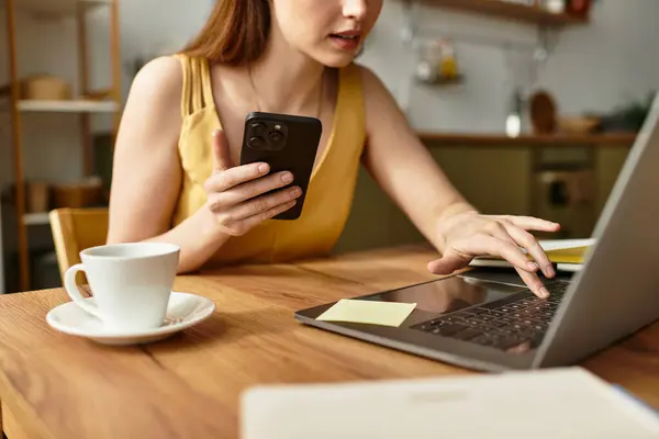 Una mujer joven se sienta en un encantador café, enfocada en su computadora portátil y sosteniendo un teléfono inteligente. - foto de stock