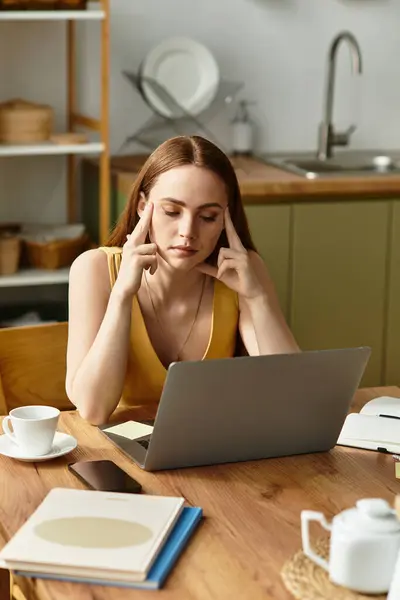 Beautiful young woman sits at a kitchen table with a laptop, deep in thought while working. — Stock Photo