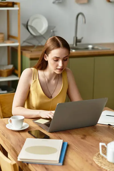 A beautiful young woman works diligently on her laptop at a wooden table in a bright kitchen. — Stock Photo