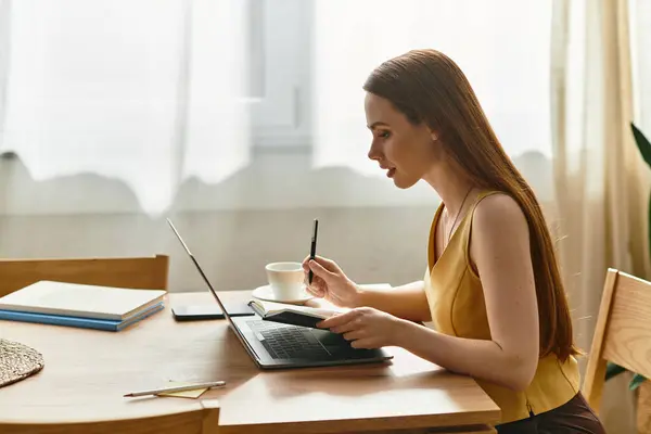 A beautiful young woman focused on her work at a sunny home office desk while sipping coffee. — Stock Photo