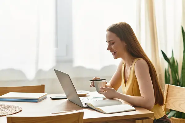 Cheerful woman sips coffee and works on her laptop at a cozy table in bright morning light. — Stock Photo