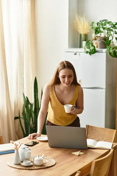 Una joven mujer sonríe a su portátil mientras toma café en un espacio luminoso y acogedor. — Stock Photo