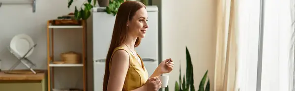Beautiful woman in a yellow dress stands near a window, sipping coffee and daydreaming. — Stock Photo