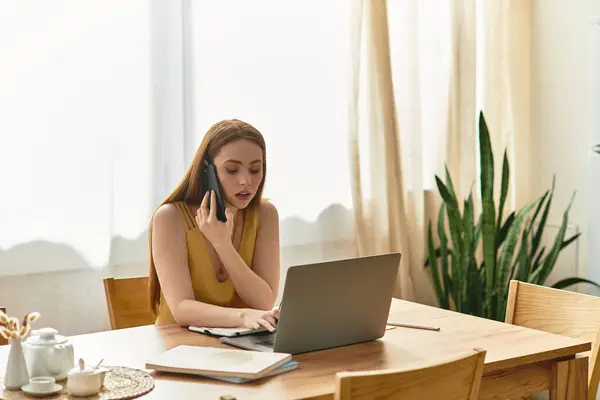 A young woman talks on the phone while working on her laptop in a sunny room. — Stock Photo