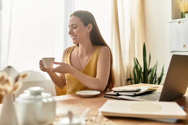 A young woman sits at a sunlit table, enjoying coffee and smiling in a cozy workspace. — Stock Photo