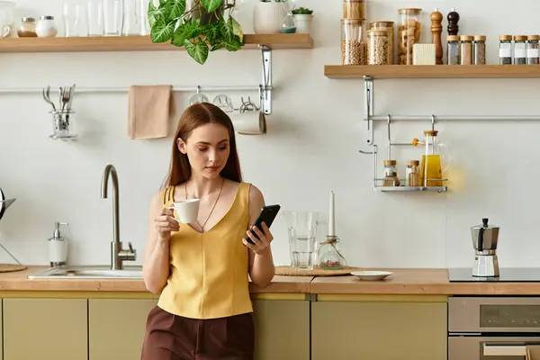A beautiful young woman stands in a bright kitchen, sipping coffee and focused on her smartphone. — Stock Photo