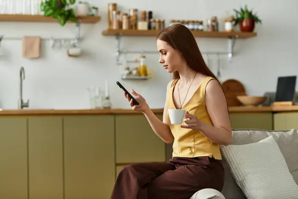 Una giovane bella donna si rilassa nella sua cucina elegante, sorseggiando caffè e usando il suo smartphone. — Stock Photo