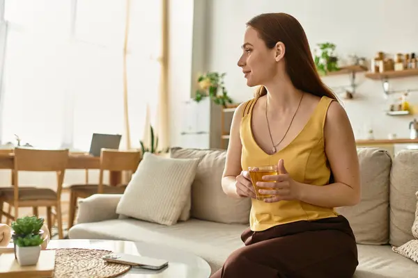 A young woman relaxes in a cozy living room, sipping a warm drink while basking in sunlight. — Stock Photo