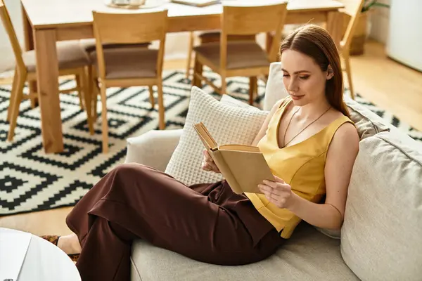 A young beautiful woman in a casual outfit reads a book while lounging on a comfy sofa. — Stock Photo