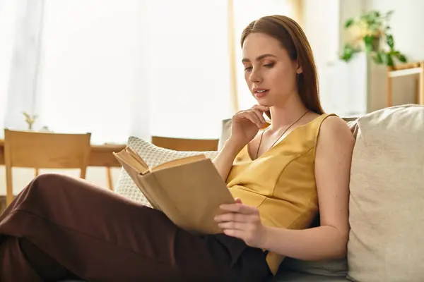 A beautiful young woman relaxes with a book on a comfy couch in a sunlit room. — Stock Photo