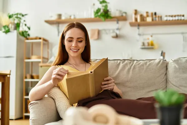 Une jeune femme aux cheveux longs s'assoit confortablement sur un canapé, immergée dans un livre dans son espace douillet. — Photo de stock