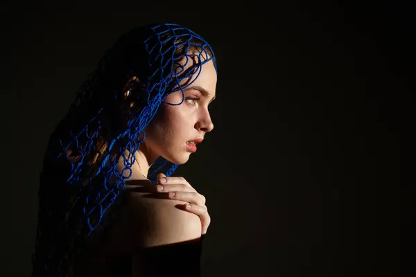 A stunning young woman wears a blue netted headpiece against a dark background, captivating all. — Stock Photo