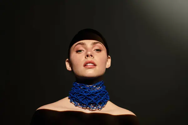 Confident young woman poses elegantly, wearing a striking blue neckpiece in a spotlight. — Stock Photo