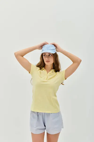 A beautiful young woman stands confidently, adjusting her cap in a soft, bright studio setting. — Stock Photo