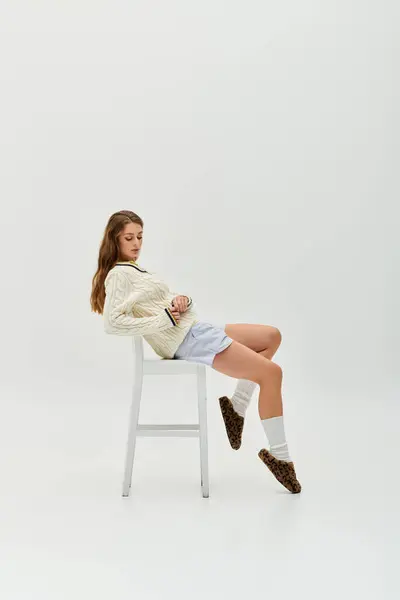 A young woman in a cozy sweater and shorts relaxes on a chair in a bright room. — Stock Photo