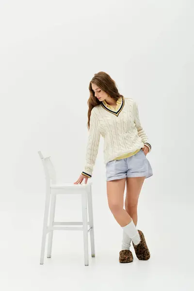 Young woman in a knit sweater and shorts playfully interacts with a white chair in a bright room. — Stock Photo